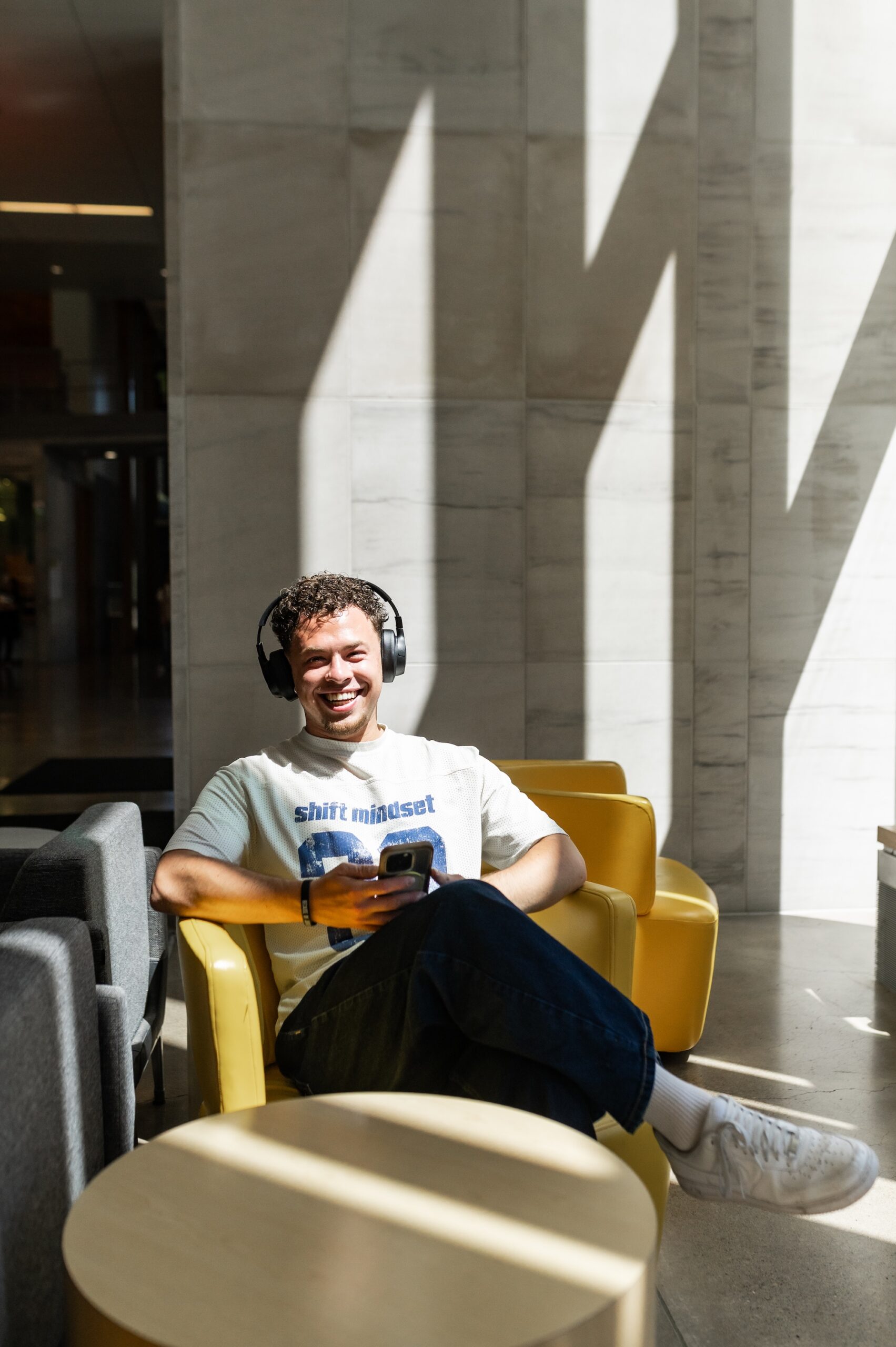 A male student wearing headphones and holding a smartphone smiling while sitting in a yellow armchair on campus scaled