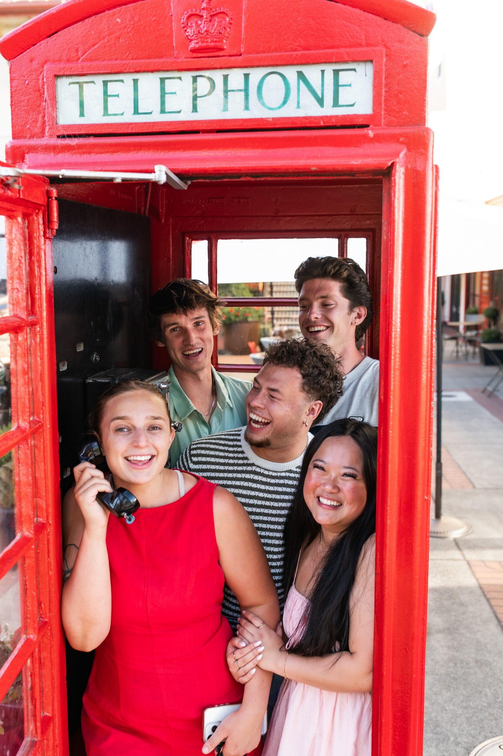 Group of five friends smiling and laughing together inside a red vintage phone booth scaled
