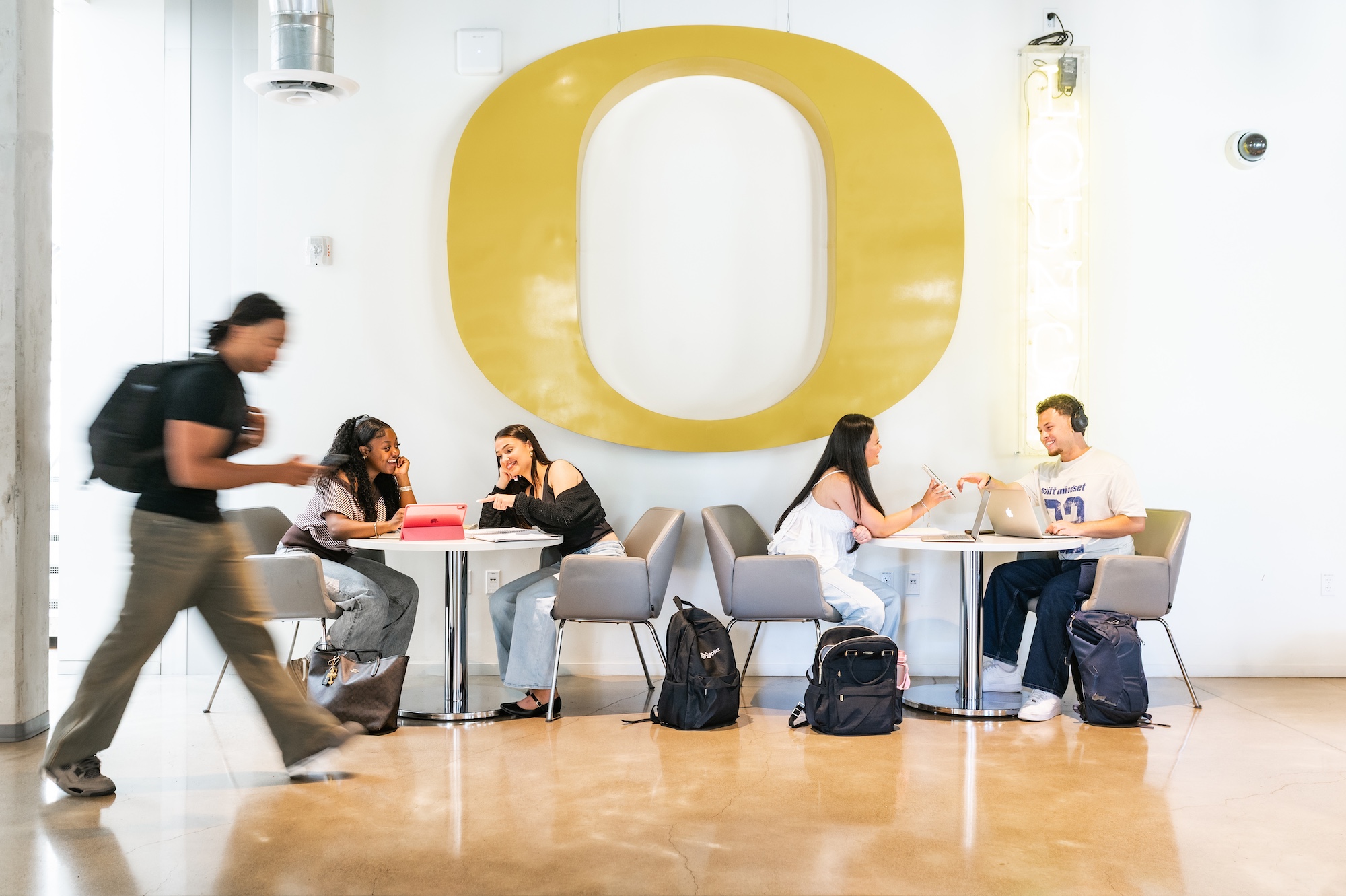 Students studying at round tables with laptops and notebooks under a large yellow University of Oregon 22O22 logo on a white wall