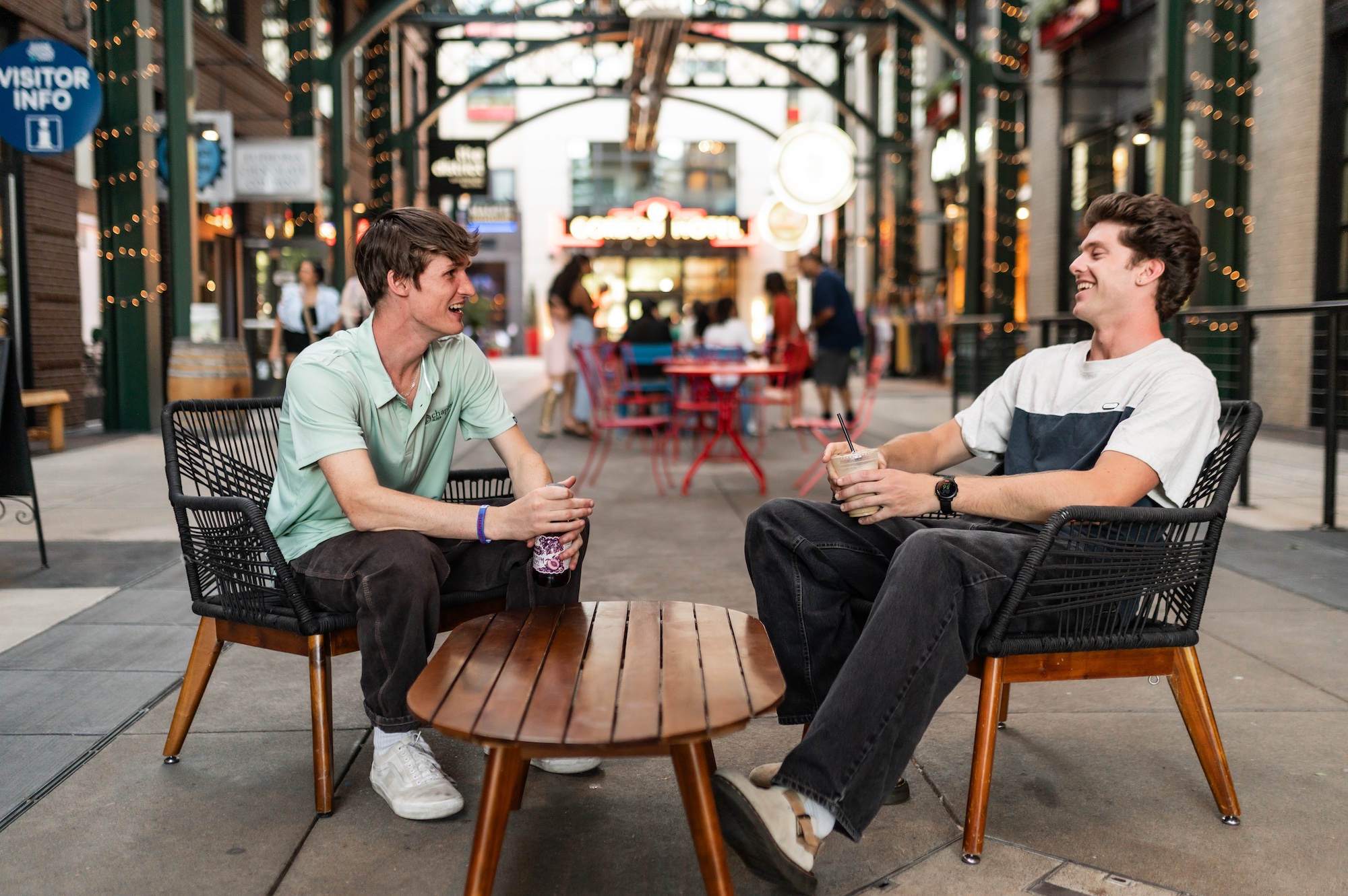 Two young men sitting in outdoor chairs laughing and talking over iced drinks at an open air mall